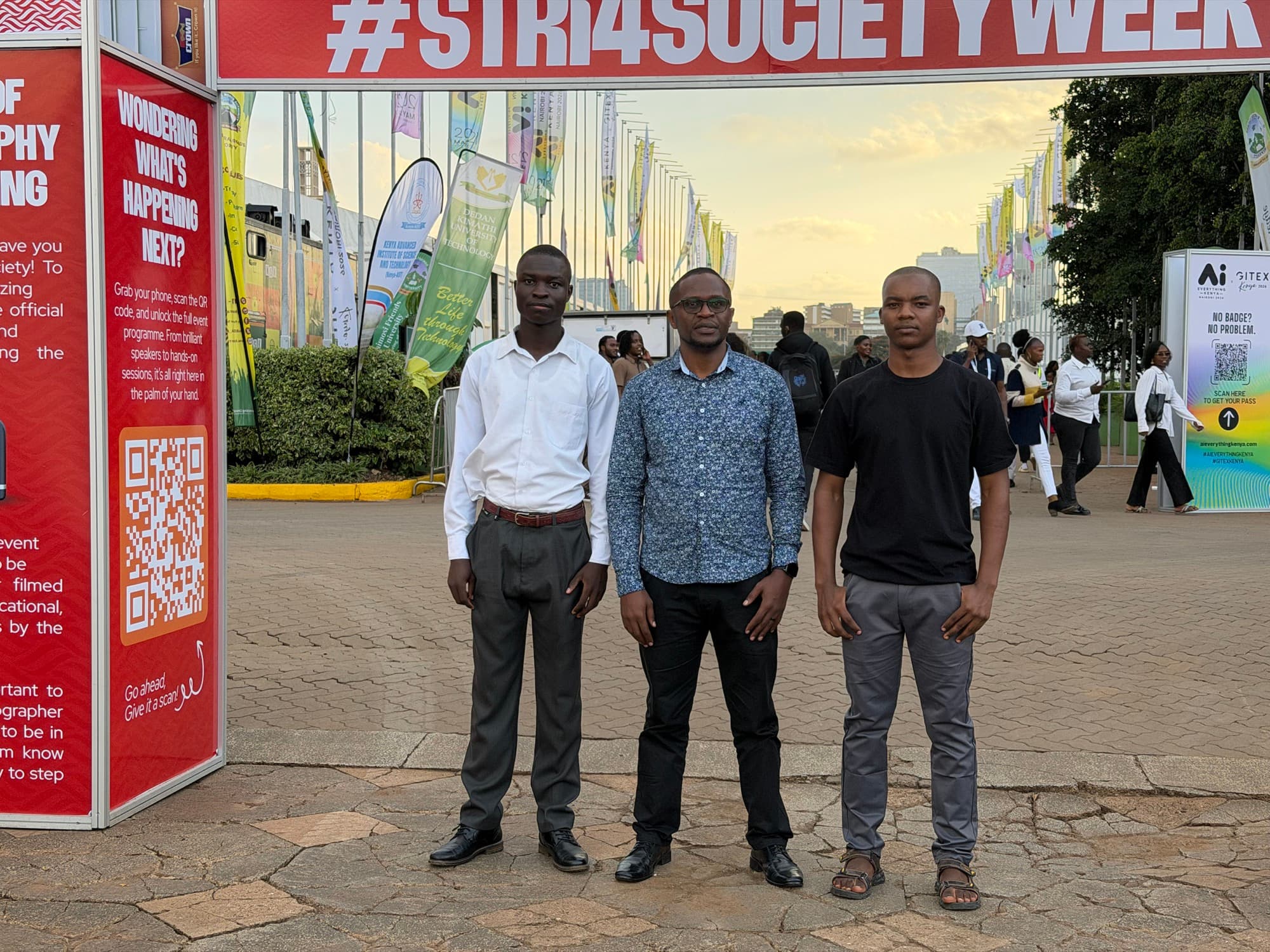 Three members of the TRACE team standing outside the KICC venue at golden hour, with #STRI4SOCIETYWEEK banners and flags lining the grounds behind them.